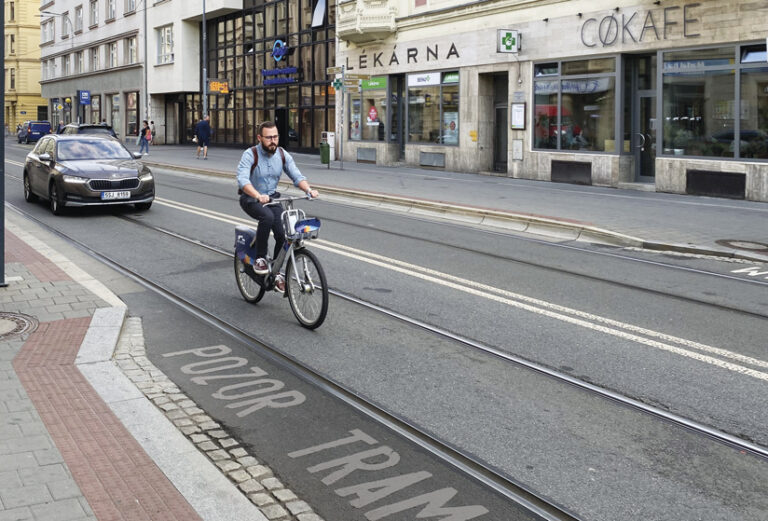 On a city street in Ostrava, a man rides a public bike-share bicycle. Streetcar tracks run through the street, and "POZOR TRAM" is painted on the asphalt. In the background are buildings and shops, including a pharmacy and a cafe. A car is also driving on the street.
