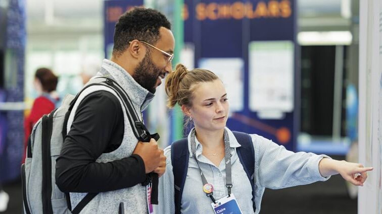 A young woman standing in front of a map with a young man, and she’s pointing to something that he’s looking at. They’re both smiling.