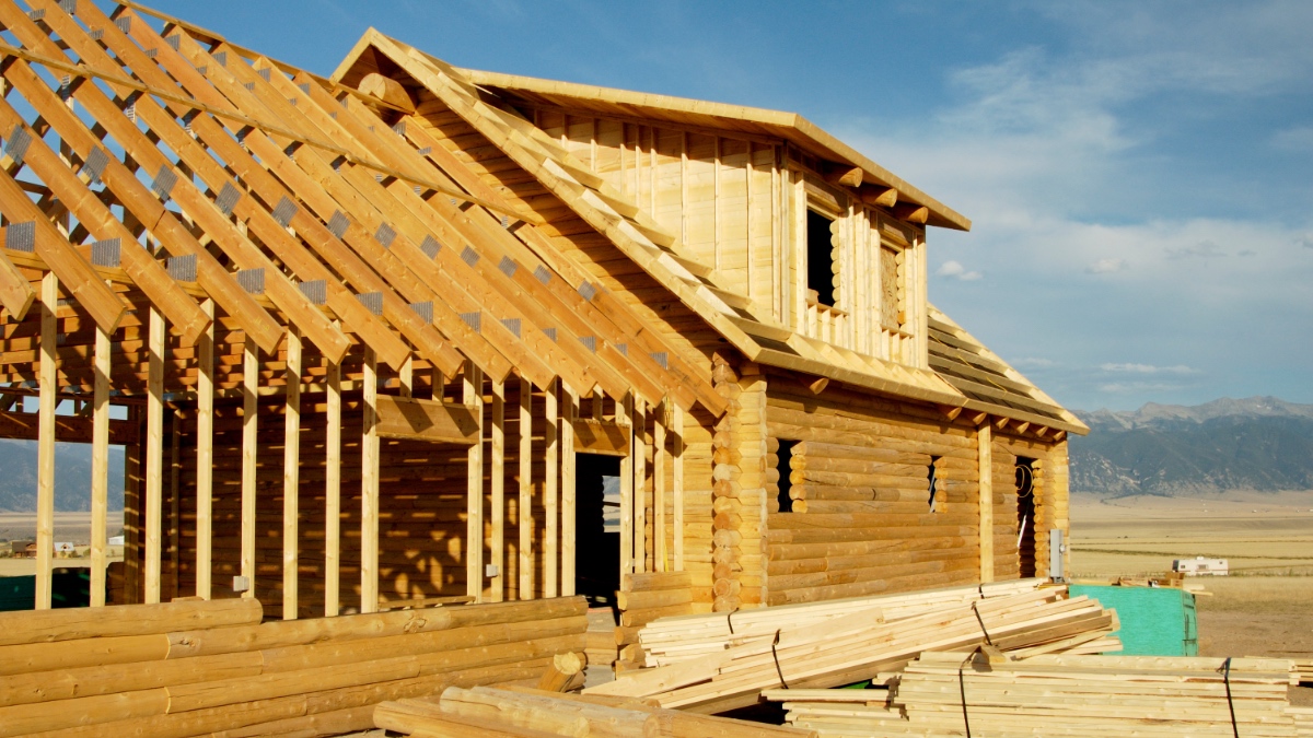 Log cabin under construction with exposed framing, set against Montana mountain valley backdrop with snow-capped peaks visible.