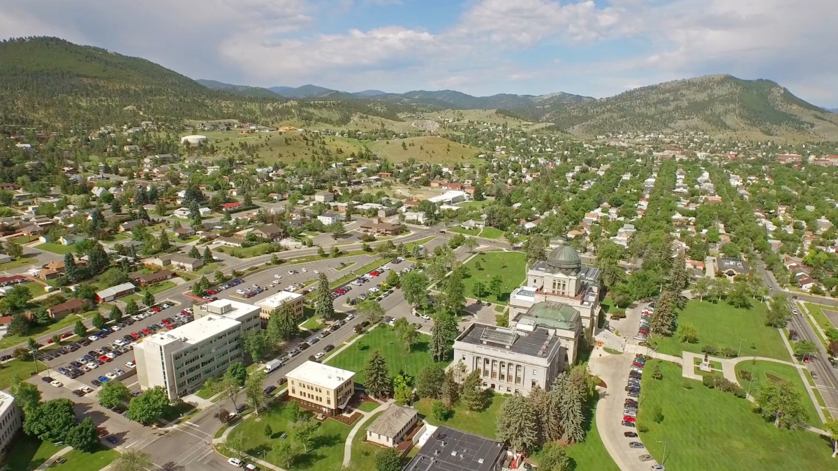 Aerial view of Montana State Capitol building with distinctive dome, surrounded by residential neighborhoods with mountains in background.