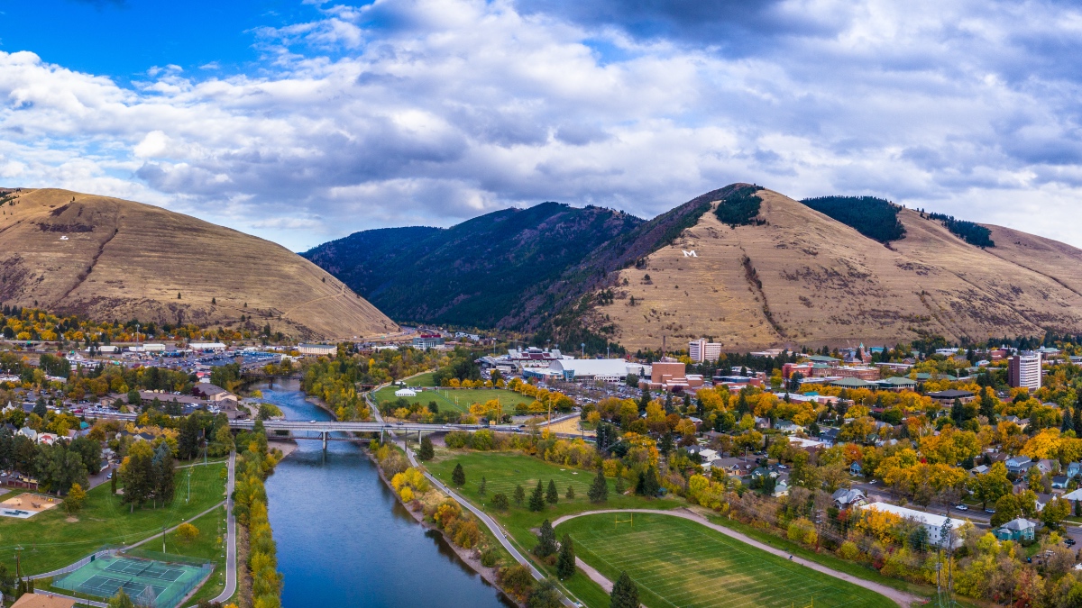 Aerial view of Missoula with Clark Fork River winding through valley, University of Montana campus visible, mountains with "M" in background.