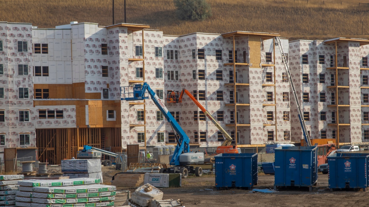 Multi-story apartment buildings under construction with construction equipment and materials in foreground, hills in background.