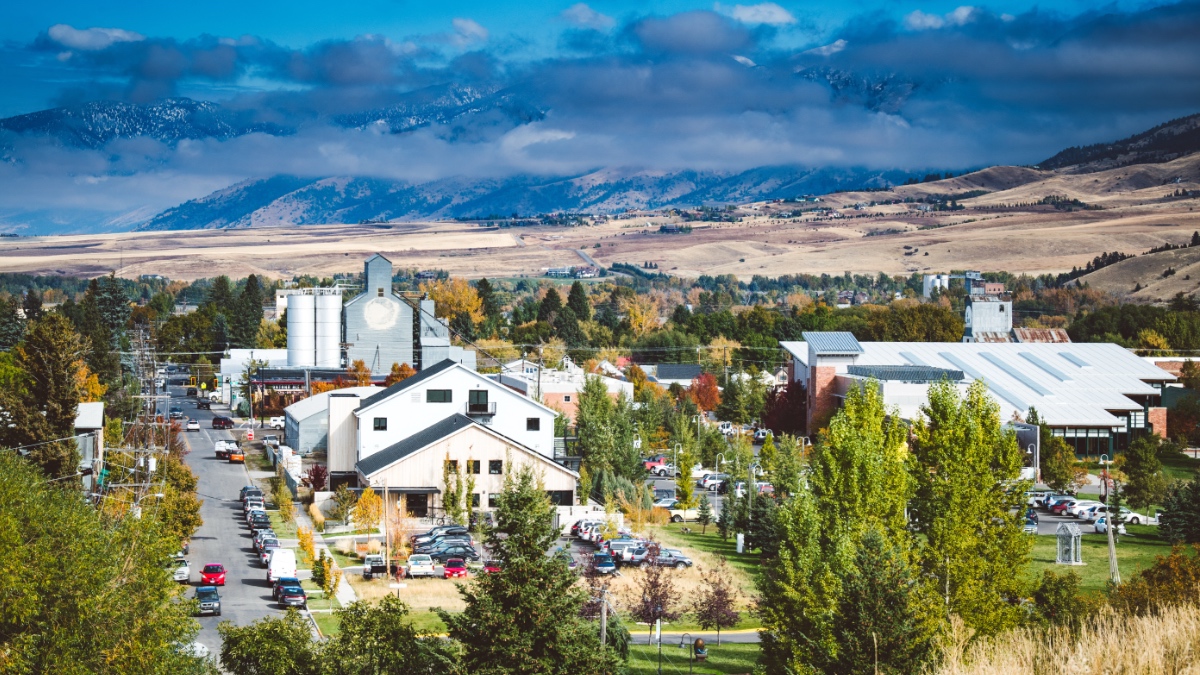 Aerial view of downtown Bozeman with mixed commercial and residential buildings, mountains and clouds visible in background.