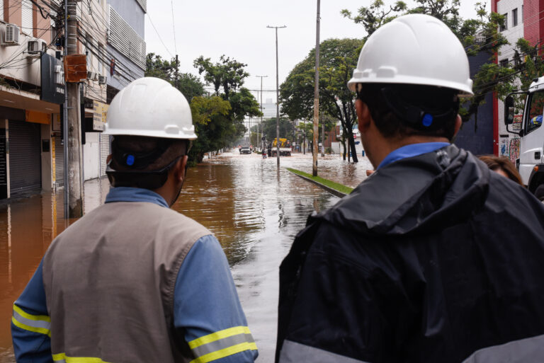 Two people in white hard hats and rain coats with their back to the viewer look down a flooded street with multi-story buildings on both sides.