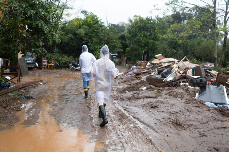 Two people in plastic tarps and rainboots walk through thick mud. Around them, debris such as broken furniture is piled high.
