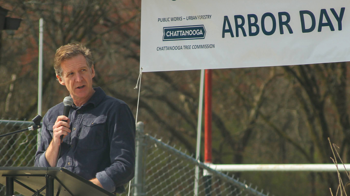 Mayor Tim Kelly speaking with microphone at outdoor Arbor Day event, with "Public Works - Urban Forestry" banner visible behind him.
