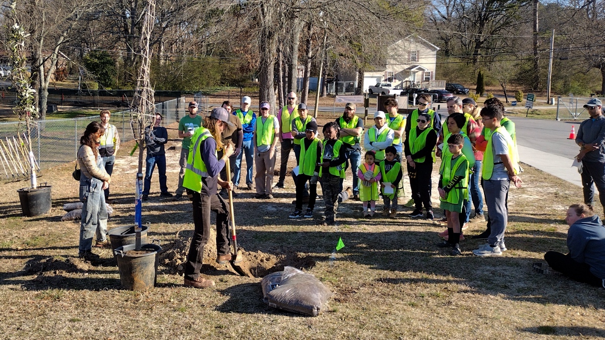 Group of volunteers in bright yellow-green safety vests gathered around instructor demonstrating tree planting technique at outdoor park site.