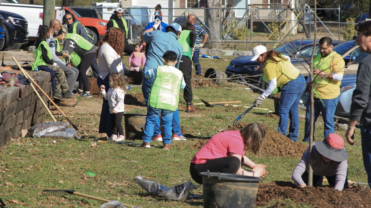 Diverse group of volunteers including adults and children working together at tree planting event, with some kneeling by fresh holes and dirt piles.