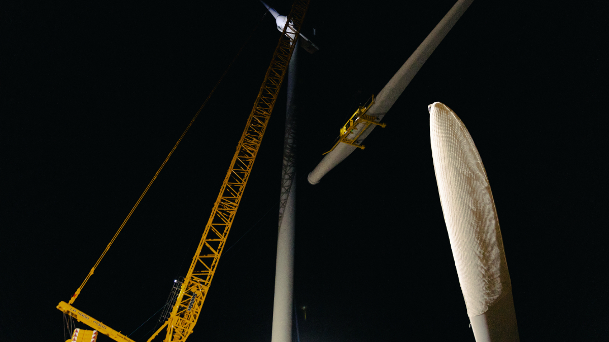 Yellow blade lift crane hoisting wind turbine components at construction site with workers and equipment on ground, turbine blade visible
