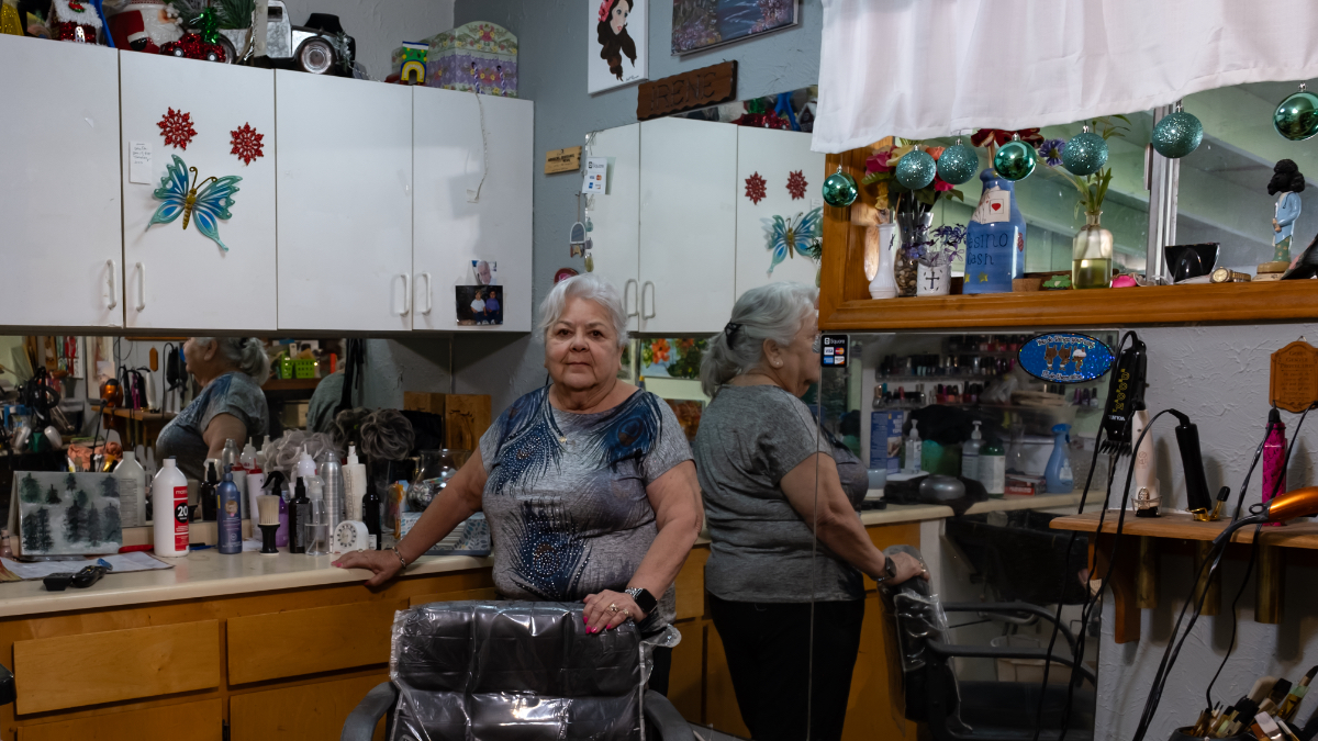 Woman stands in home salon with hair products on counter, mirrors, and cabinets in background, holding edge of counter