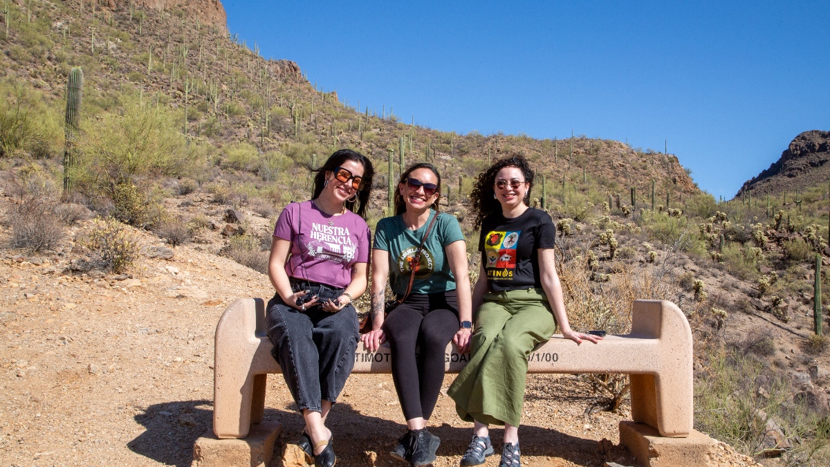 Three women stand in a desert landscape with cacti and mountains in the background, posing during a research road trip