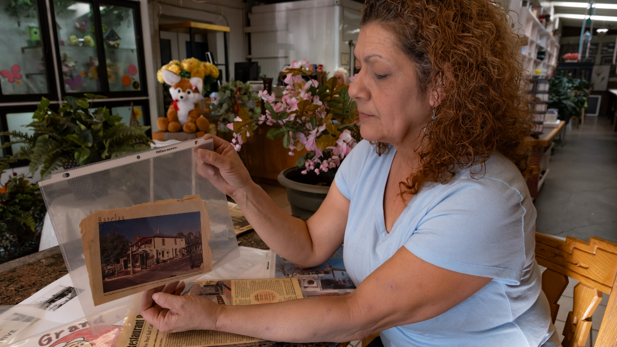 Person seated at a table in a flower shop holding a photo, surrounded by flowers, papers, and a “Grand Opening” card