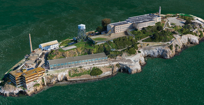 A realistic digital twin of Alcatraz Island showing rocky terrain, the main prison building, the lighthouse, and a water tower—all surrounded by green water.