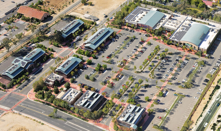 An aerial view of a modern commercial complex, as modeled in a digital twin. It shows multiple buildings, some with solar panels, encircling a large, car-filled parking lot.