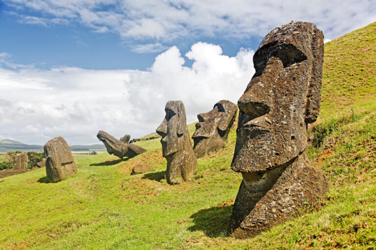 Partially buried moai statues dot a grassy hillside under a partly cloudy sky; one is in close-up profile on the right.