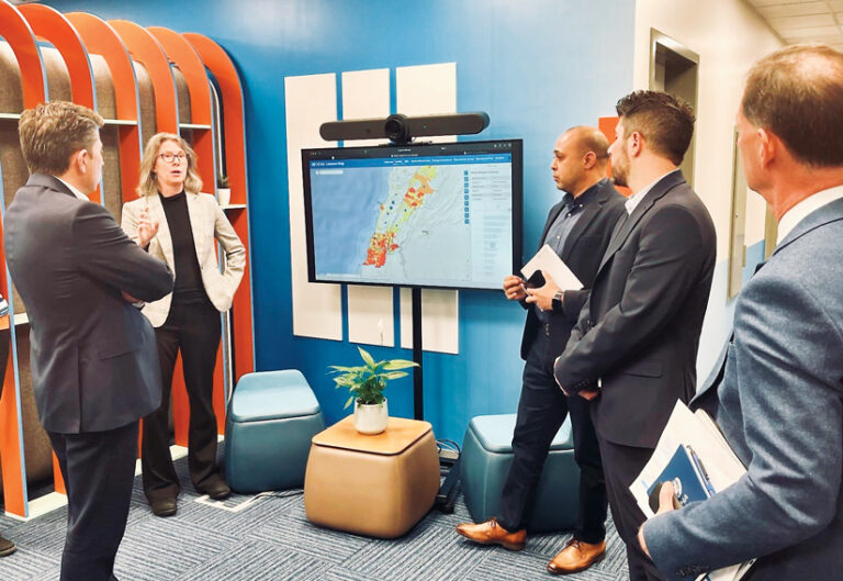 Five business-attired people present a color-coded map on a large screen in a modern office.