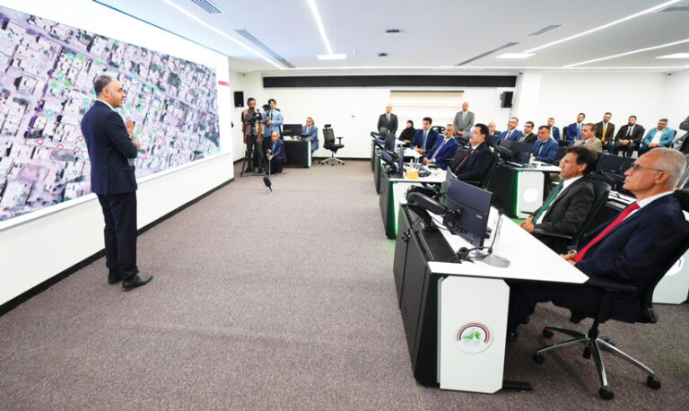 A man stands in front of a projector screen with a satellite imagery-based map on it, talking to a room full of people sitting at tables with computer screens on them.