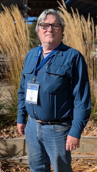 A man with gray hair and glasses wearing jeans, a navy blue button-up shirt, and a conference badge stands in front of tall grassy plants.