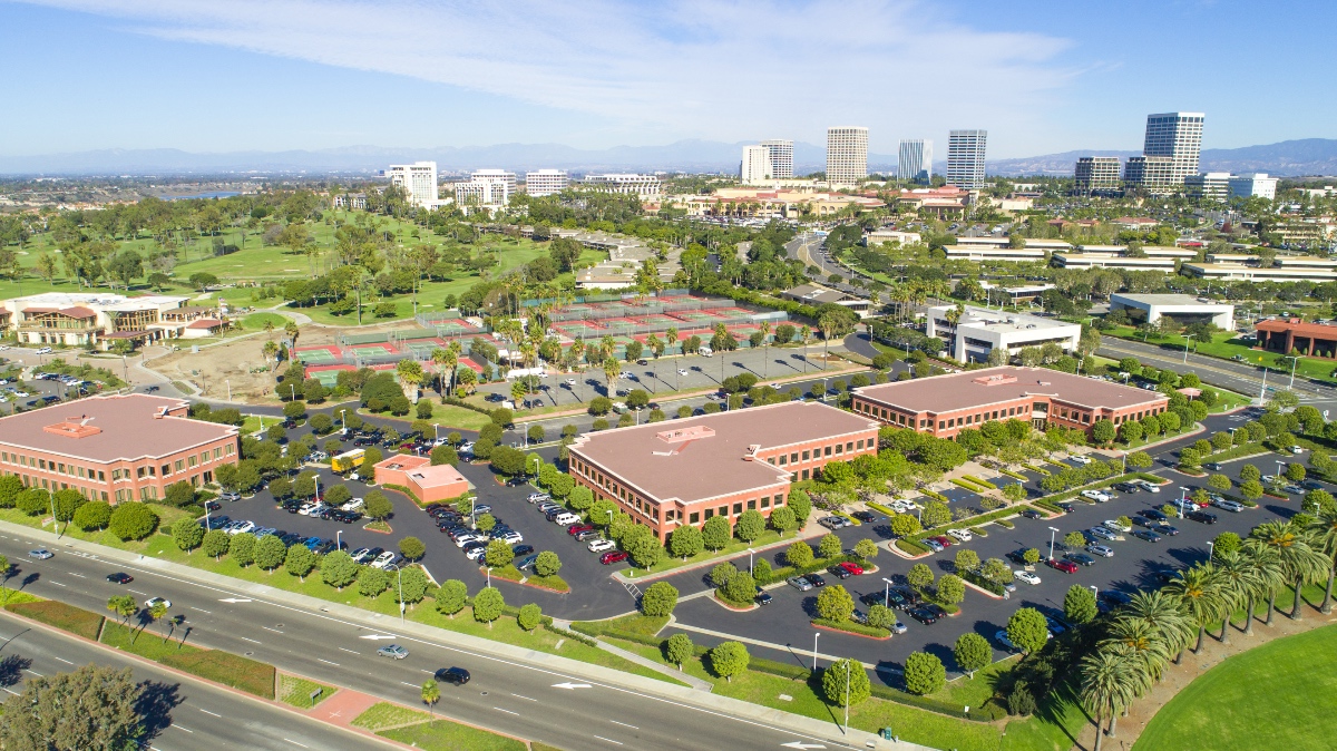 Aerial view of Irvine, California showing office buildings surrounded by tennis courts, green lawns, tree-lined streets, and expansive park areas, with downtown high-rises and mountains in the distance