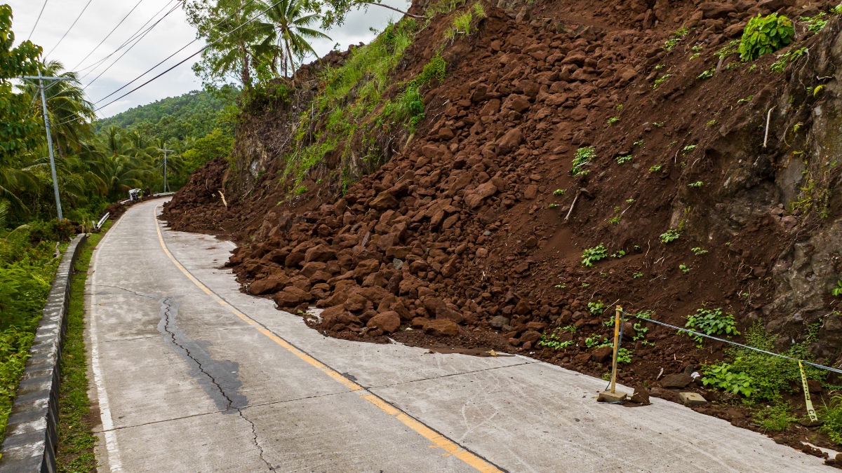 Large landslide of rocks and soil blocking a curved mountain road in the Philippines, with yellow caution tape and forested hills in background.