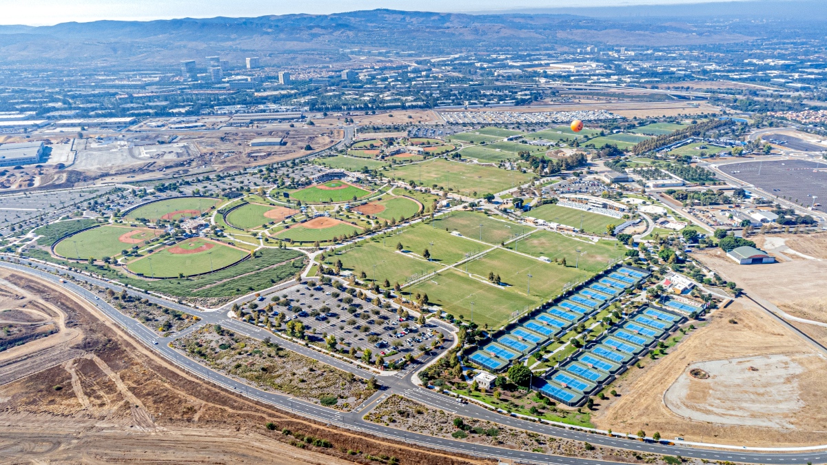 Aerial photograph of Irvine, California showing extensive green spaces including baseball diamonds, soccer fields, tennis courts, and parks woven throughout residential development with mountains visible in the background