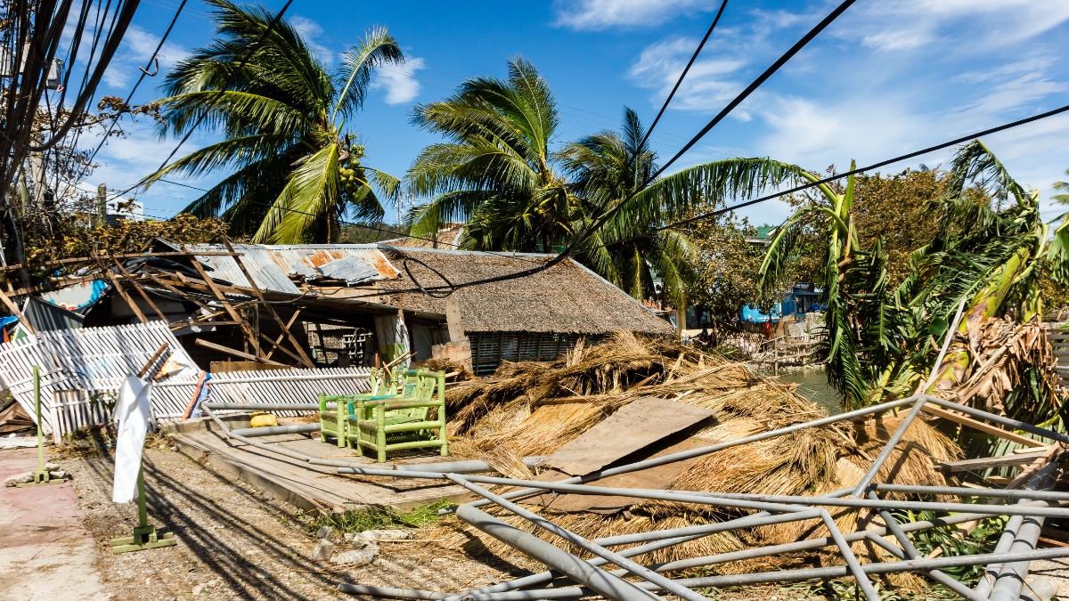 Typhoon-damaged Philippine village with collapsed thatched-roof homes, torn corrugated metal, downed power lines, and debris scattered among damaged palm trees.