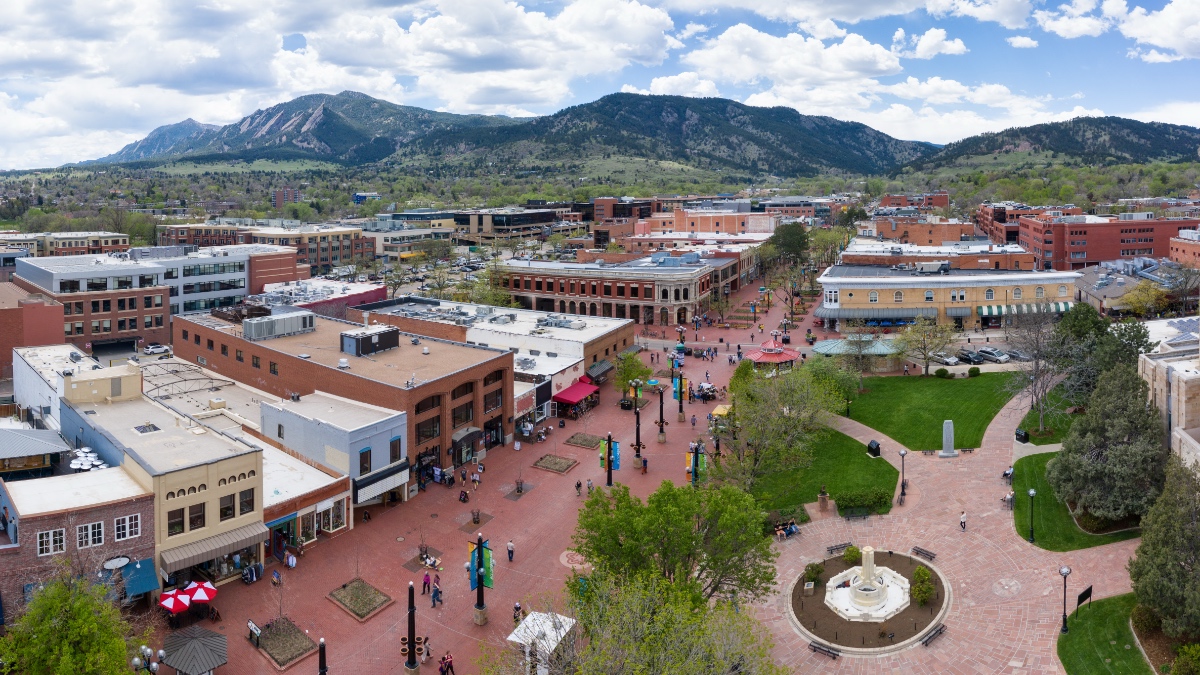 Aerial view of Boulder's Pearl Street pedestrian mall with red brick plaza, outdoor vendors, fountain, and green spaces, backed by the Flatiron mountains and foothills
