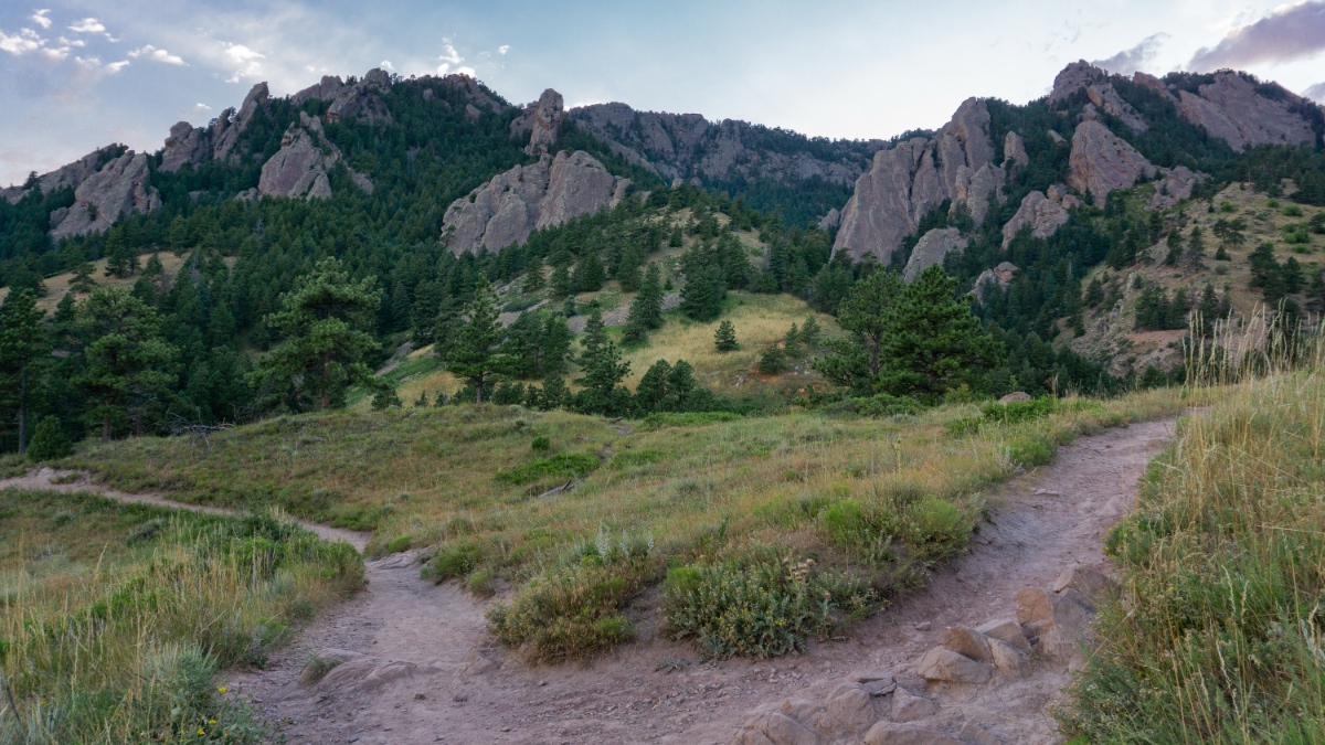 Hiking trails wind through grassland toward the Flatirons rock formations near Boulder, Colorado, with forested slopes and dramatic sandstone slabs rising against a cloudy sky