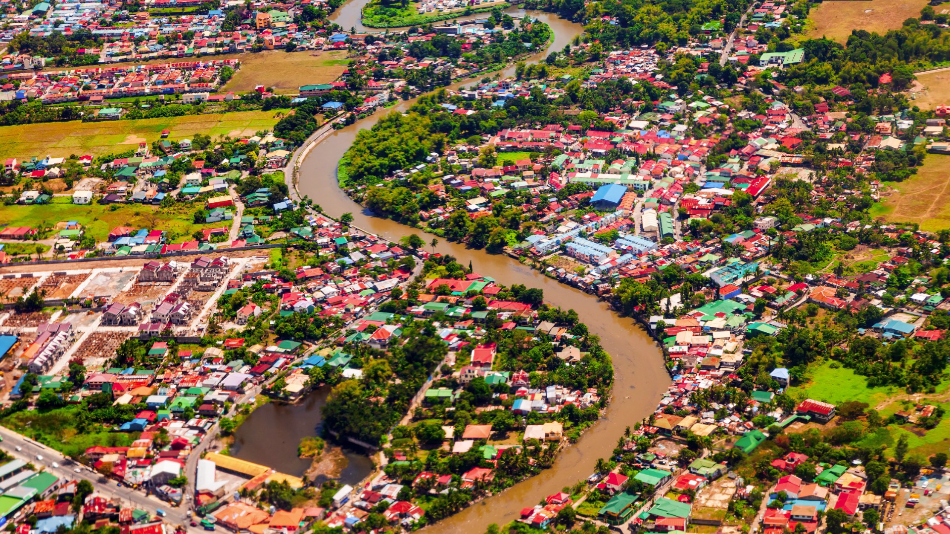 Aerial view of Manila showing winding brown river cutting through dense residential neighborhoods with colorful rooftops, green spaces, and farmland.