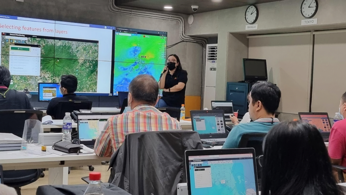 Trainer leading HANDA GIS workshop with participants at laptops, large screens displaying hazard maps and layer selection tools, clocks showing Manila and Tokyo time.