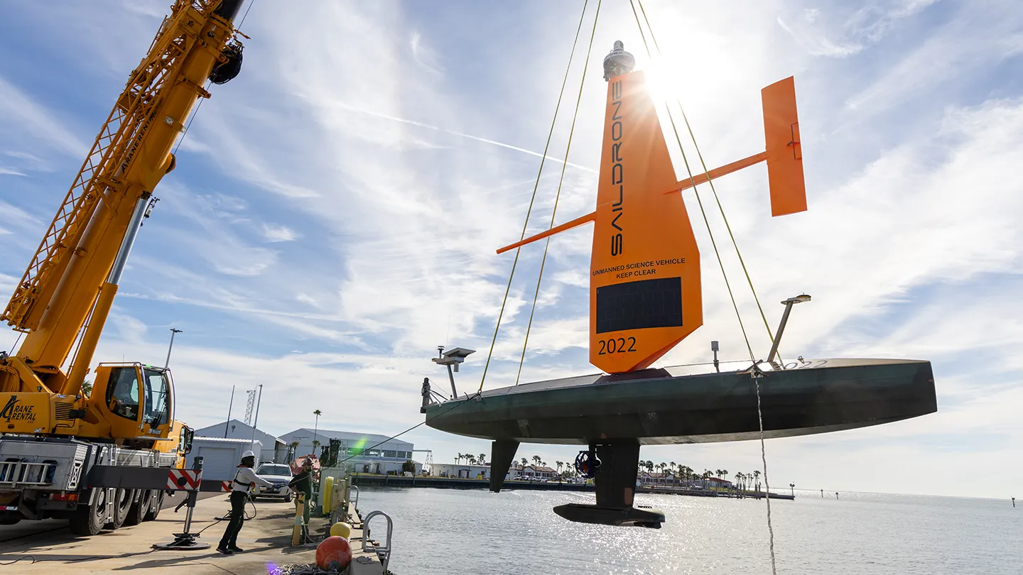 Orange autonomous Saildrone vessel with rigid wing sail being lifted by crane over water during launch, with palm trees visible in background.