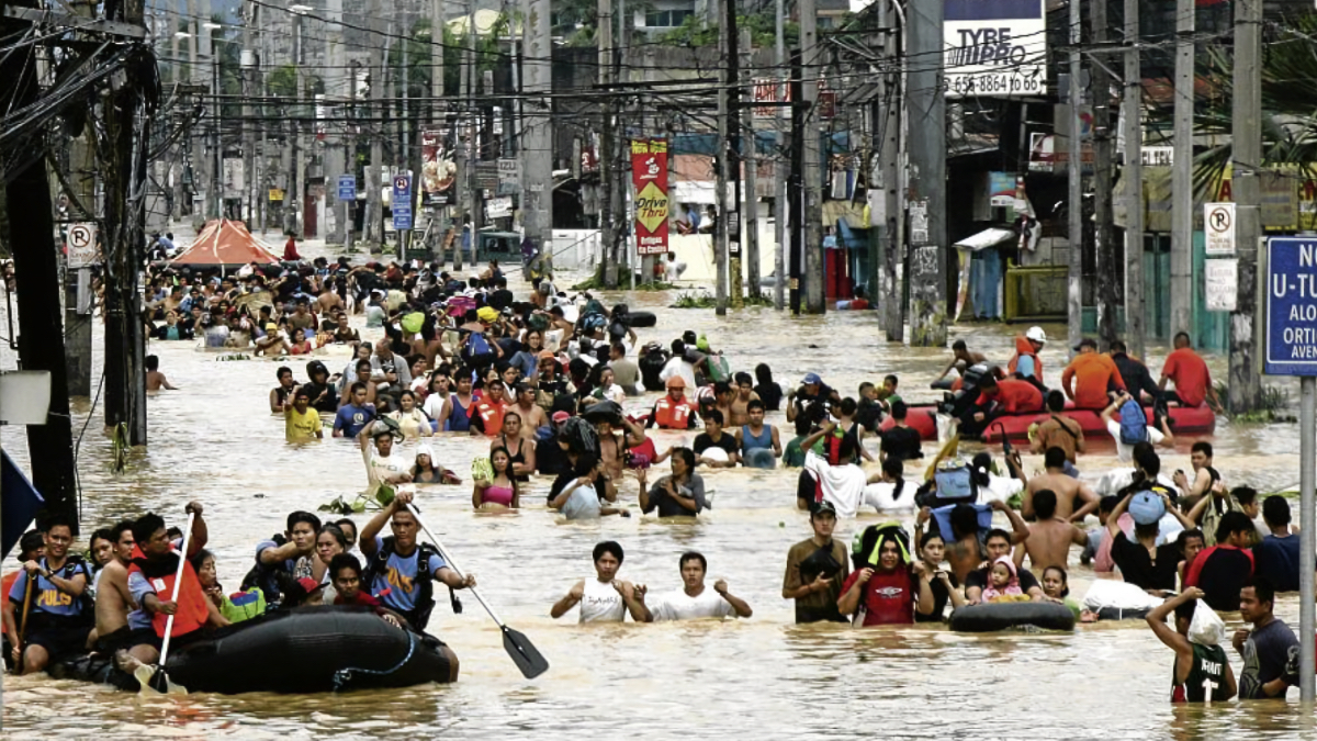 Crowds of people wading through chest-deep floodwater on urban Manila street during Typhoon Ketsana, with power lines overhead and rescue boats visible.