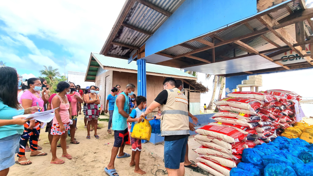 Relief coordinator in vest distributing supplies to masked families waiting in line, with stacks of rice bags and aid packages under shelter after typhoon.