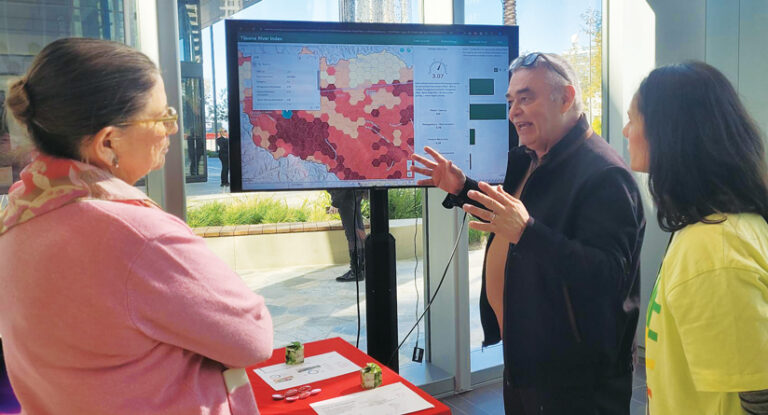 A man gestures at a large screen displaying a color-coded map, presenting to two women, one in pink, the other in yellow.