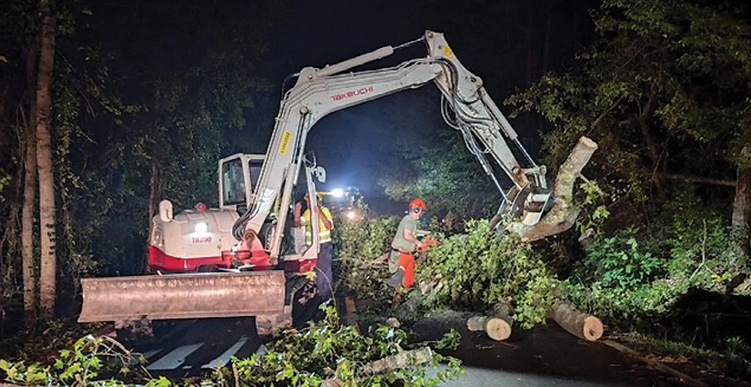At night, a white excavator lifts a large tree trunk as workers clear fallen branches from a road with chainsaws.