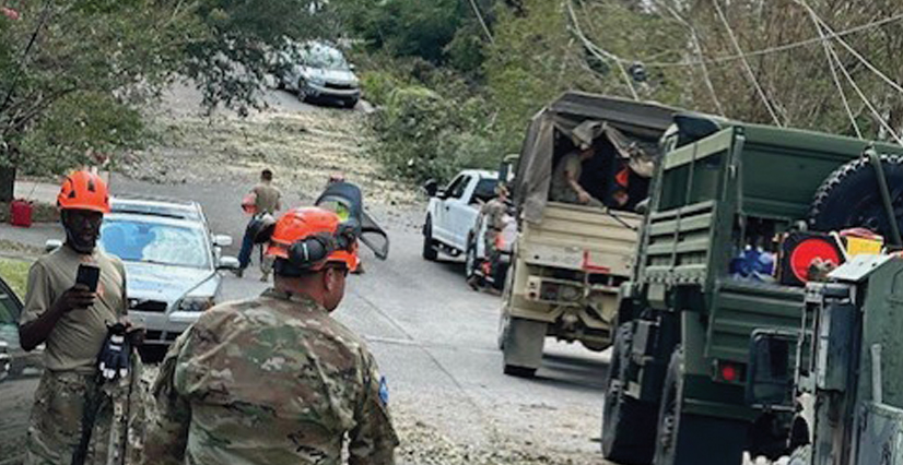 Military personnel in camouflage and orange hard hats clear a debris-strewn street with a large military truck present.