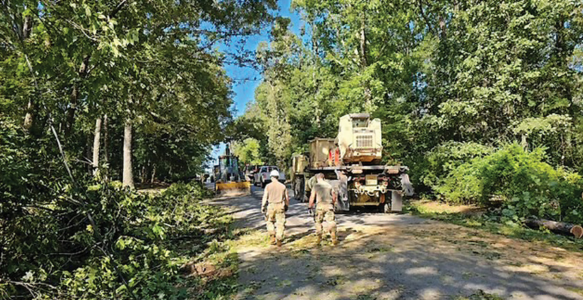 Two soldiers in camouflage walk on a wooded road where heavy machinery clears fallen trees.