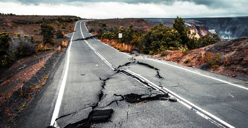 A cracked, buckled paved road running through volcanic landscape with a smoking crater in distance.
