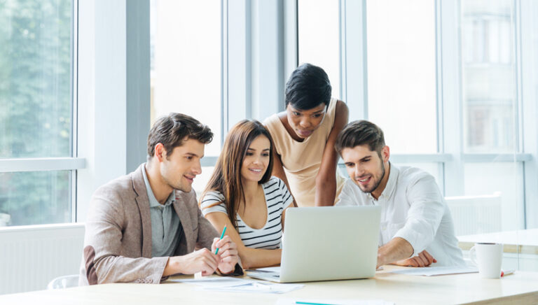 Four young people in a bright office setting gather around a laptop, looking and pointing at the screen.