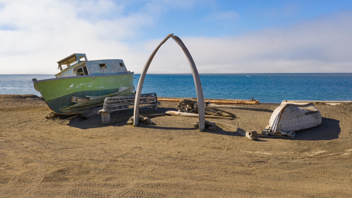 Whale bones form an arch on an Arctic Alaska beach beside a weathered boat and traditional watercraft, with the open sea stretching to the horizon.