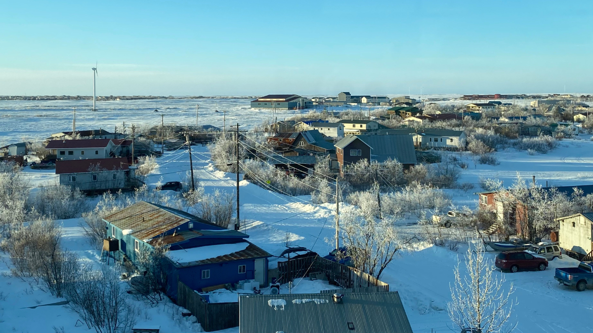 Aerial view of Bethel, Alaska in winter, with snow-covered homes, frost-lined trees, and flat tundra extending to the horizon beneath a clear blue sky.