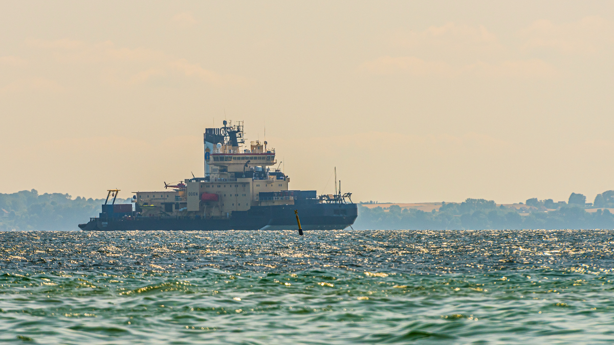 The Swedish icebreaker Oden, a large dark-hulled research vessel with a multi-story white superstructure, sits on calm sunlit waters with a hazy coastline in the background.