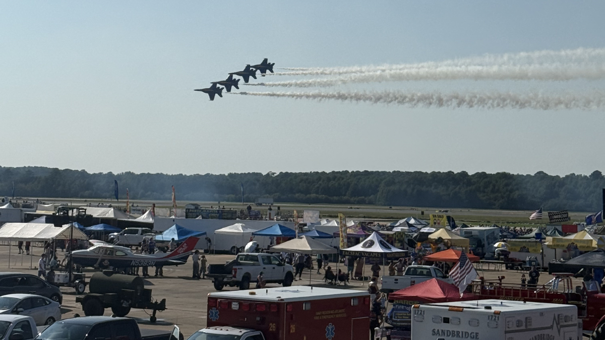 Blue Angels flying in formation over Navy air show grounds with crowds, tents, emergency vehicles, and aircraft displays below.