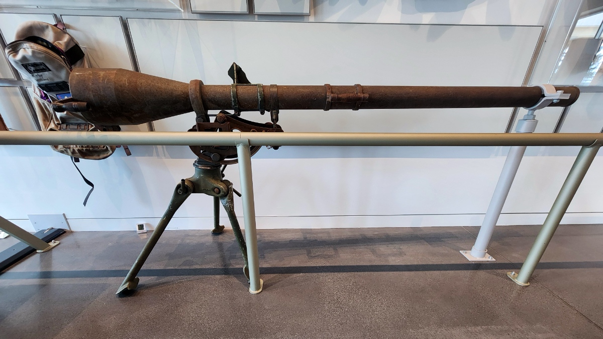 Vintage avalanche control cannon mounted on tripod, displayed indoors with weathered steel barrel and mechanical sighting equipment.
