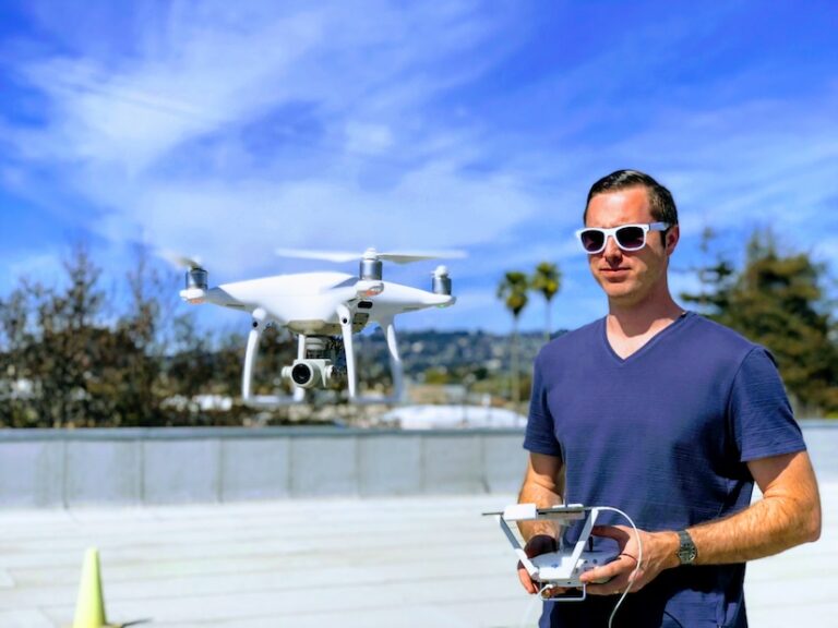 A young man wearing white sunglasses holding a remote and flying a drone