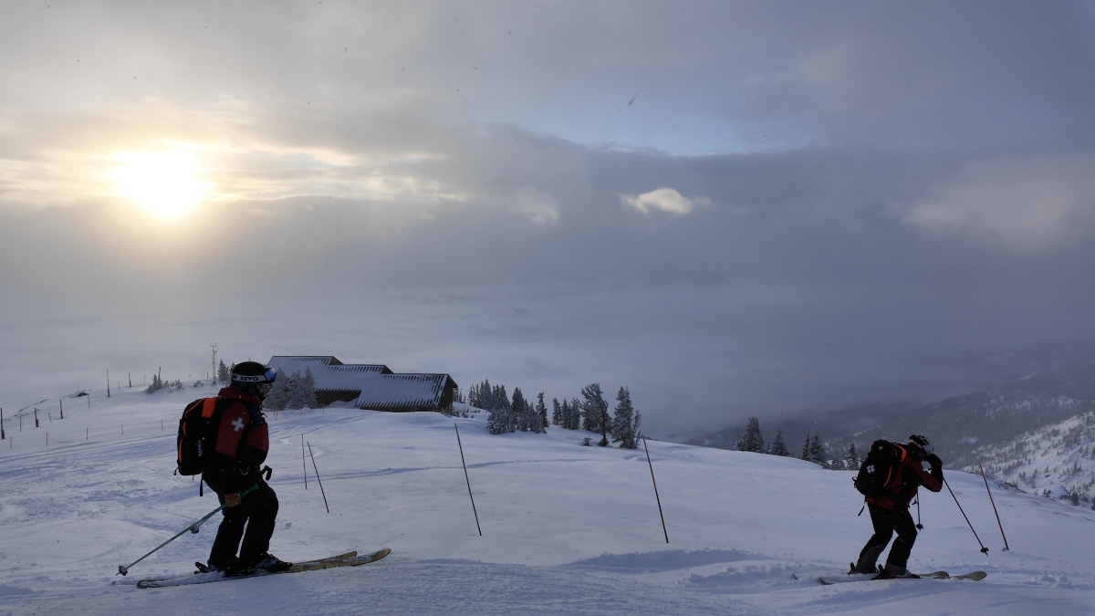 Two ski patrollers in red jackets ski toward mountain buildings at dawn, pre-sunrise blue light illuminating snow-covered slopes and valley below.