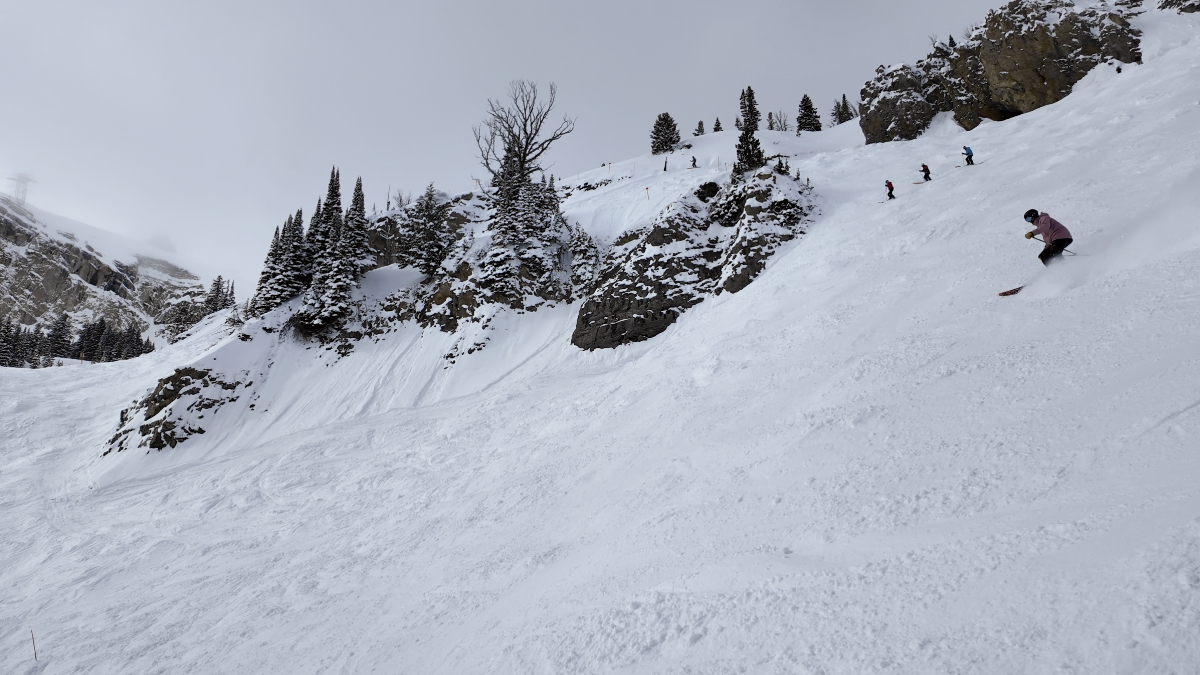 Multiple skiers descending steep slope with exposed rock cliffs and evergreen trees, fresh snow tracks visible across the face.