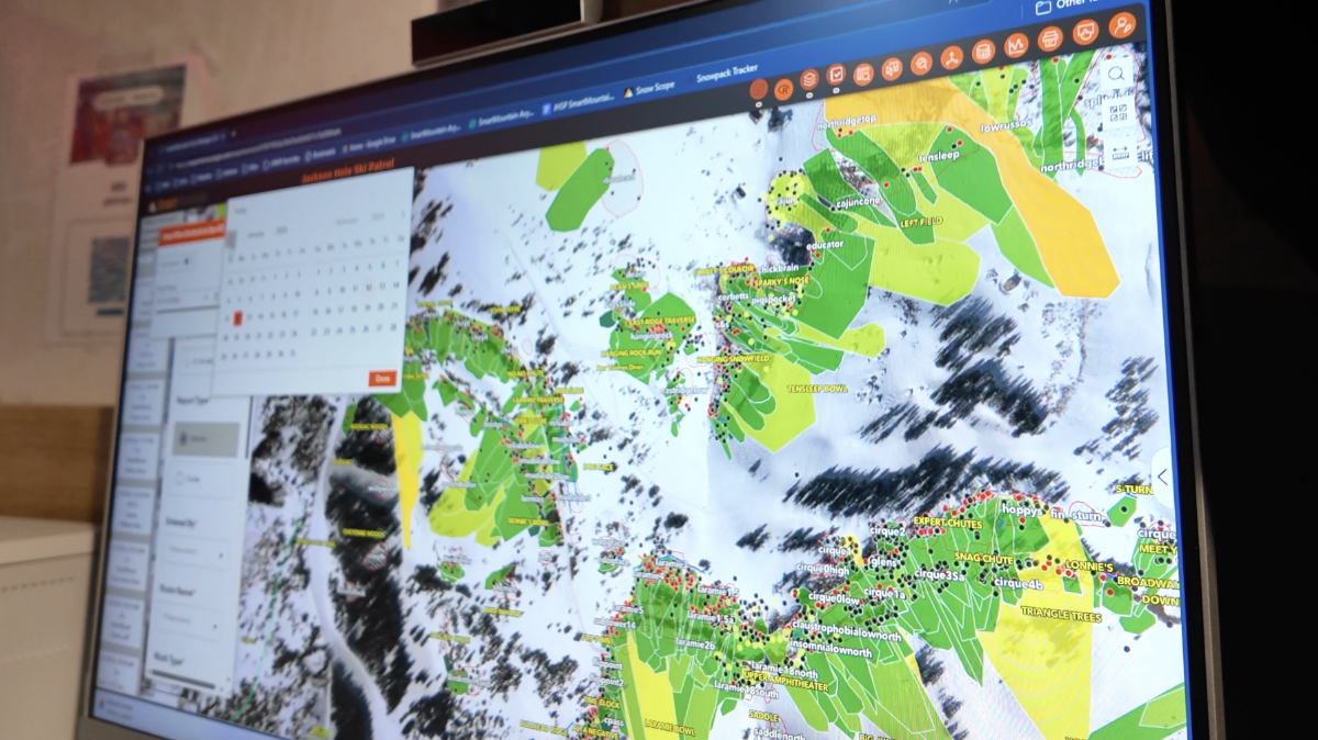 Computer monitor showing GIS map with green and yellow polygons overlaid on aerial imagery of snowy mountain terrain with multiple colored dots.