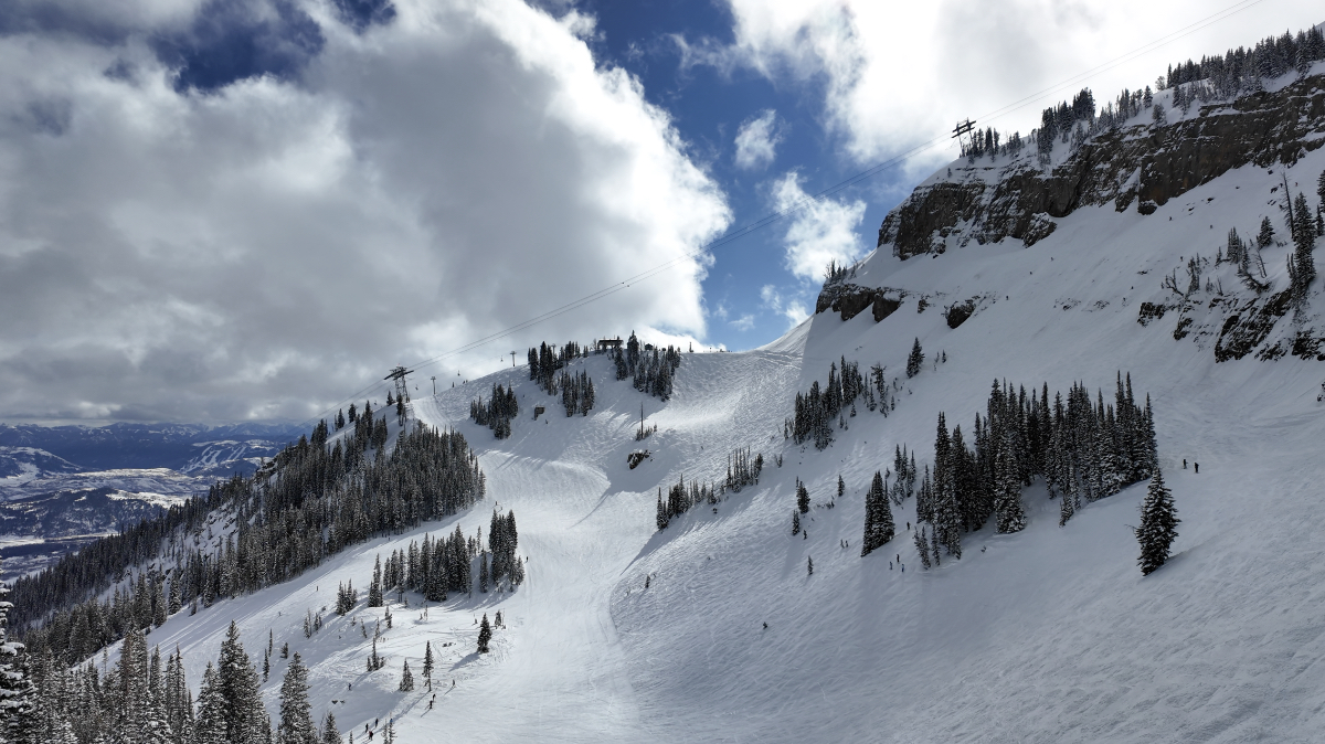 Wide view of Jackson Hole ski resort showing tram cable line crossing steep terrain with rock cliffs, scattered evergreens, and skiers on slopes.