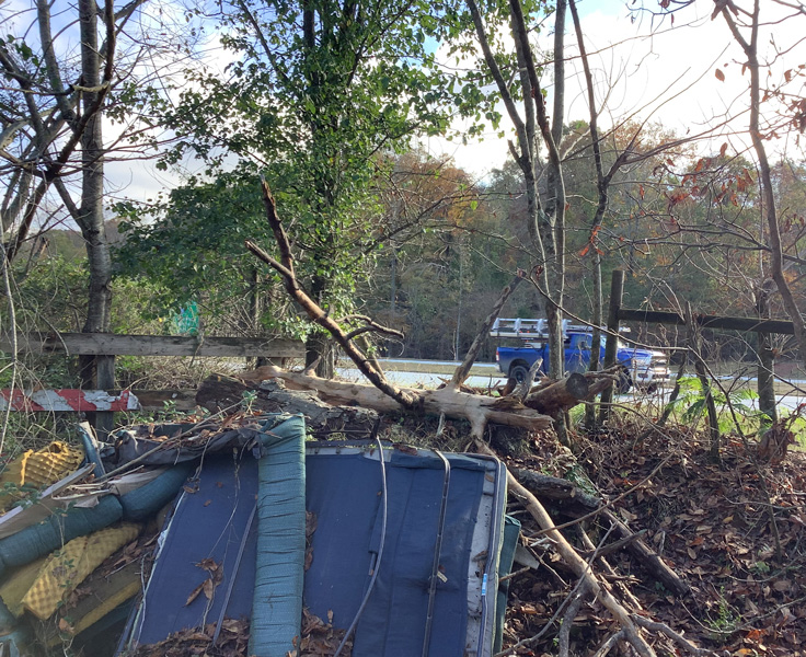 Photo of the side of a road where various kinds of trash have been dumped, including a discarded mattress and sheets of yellow foam. A blue truck drives by on the road in the background of the image.